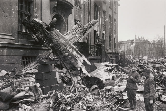 Black and white image of aircraft wreckage against a damaged building with two soldiers observing.