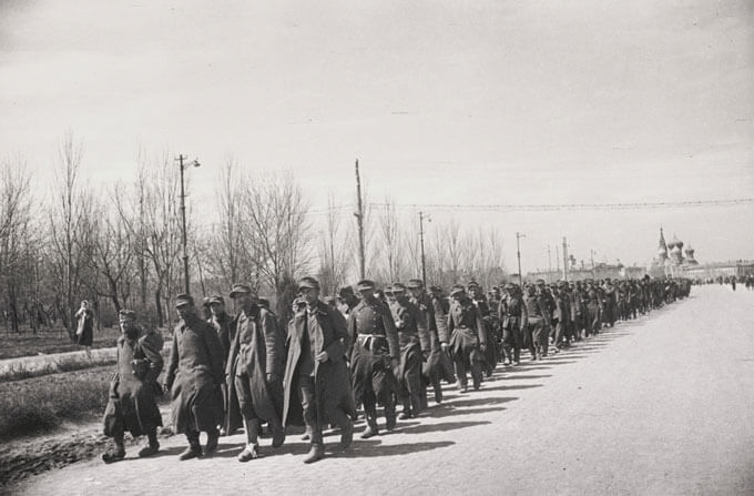A group of soldiers in uniform marching along a road with bare trees and a church in the distance.