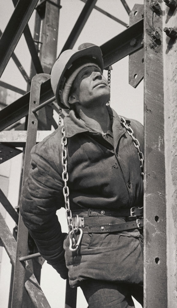 A black and white photo of a man wearing a construction helmet standing on a metal framework and gazing upwards.