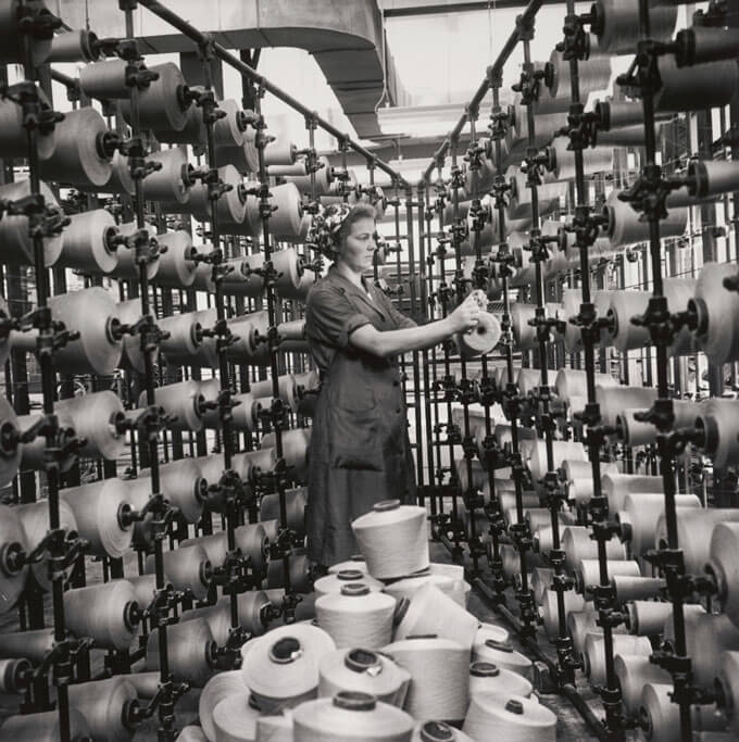 A black and white photo of a person collecting spools of thread in an industrial setting.