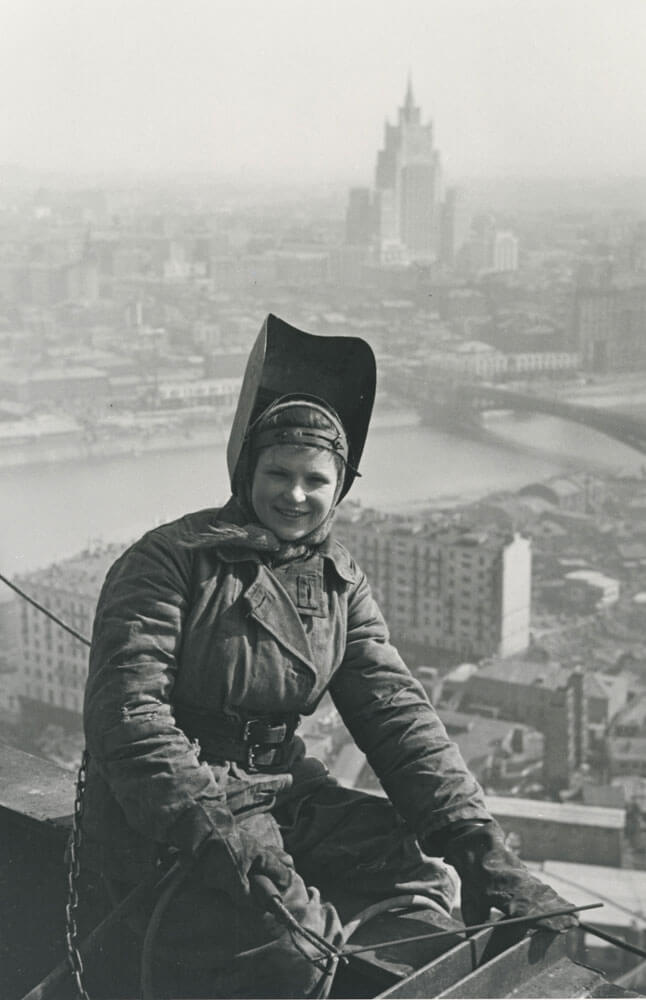 A black and white photo of a person sitting atop a girder elevated in the air with a skyscraper in the background.