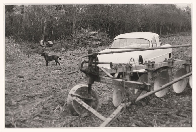 Car parked next to a plow in a field. Two men kneel over the ground in the background, with a dog standing in the foreground.